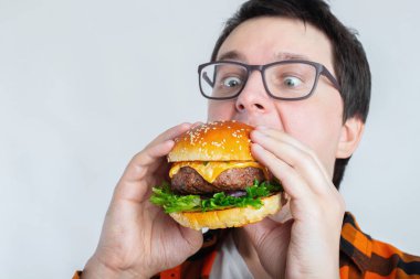 Taze bir Burger tutan gözlüklü genç bir adam. Çok aç bir öğrenci fast food yiyor. Sıcak yararlı yemek. Oburluk ve sağlıksız beslenme kavramı. Kopya alanı ile metin için.