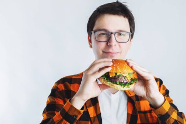 Taze bir Burger tutan gözlüklü genç bir adam. Çok aç bir öğrenci fast food yiyor. Sıcak yararlı yemek. Oburluk ve sağlıksız beslenme kavramı. Kopya alanı ile metin için.
