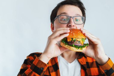 Taze bir Burger tutan gözlüklü genç bir adam. Çok aç bir öğrenci fast food yiyor. Sıcak yararlı yemek. Oburluk ve sağlıksız beslenme kavramı. Kopya alanı ile metin için.