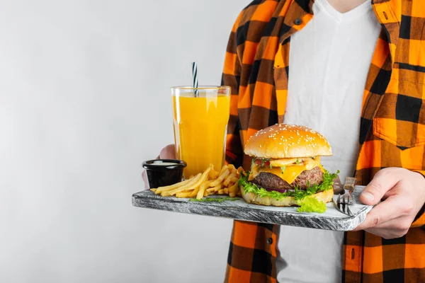 A male student in a checkered orange shirt on a white background is holding a wooden Board with a fresh Burger, a glass of orange juice and fried potatoes. With copy space for text