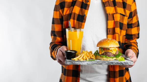 Un estudiante masculino con una camisa naranja a cuadros sobre un fondo blanco sostiene una mesa de madera con una hamburguesa fresca, un vaso de jugo de naranja y papas fritas. Con espacio de copia para texto — Foto de Stock