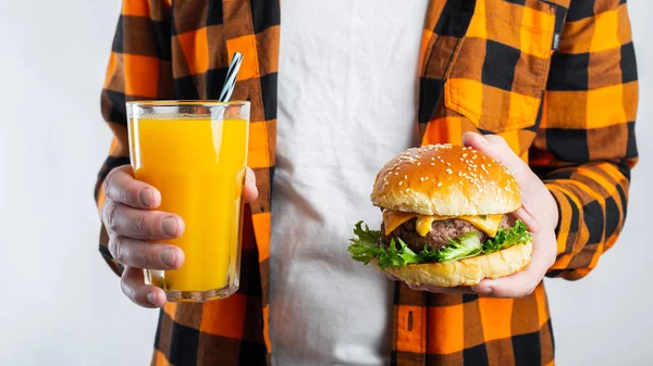 Un estudiante masculino con una camisa naranja a cuadros sobre un fondo blanco sostiene una hamburguesa fresca y un vaso de jugo de naranja. El concepto de alimentación poco saludable entre los escolares — Foto de Stock