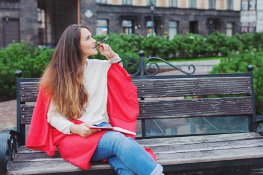 Uzun kahverengi saçlı çekici bir kız kırmızı halı gizleme, bir kalem kemiren ve kentsel bir arka plan üzerinde düşünme bir bankta oturur. O onun elinde bir defter vardır.