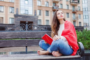 Çekici kız çıplak ayaklı bir bankta oturan yeni yerleşim bölgesinde kırmızı bir battaniye ile örtülü ve düşüncelerini içine kırmızı bir defter yazıyor. Kentsel arka plan.