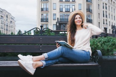 Şık bir şapka bir genç güzel kadın yeni bir konut mahallede bir bankta oturur ve bir kağıt kitap okur. O sayfaları ve gülümseme ile çevirir. Kentsel arka plan.