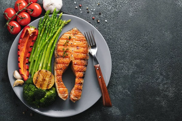 Kuşkonmaz, brokoli ve gri bir plaka üzerinde kırmızı biber ile somon steak lezzetli ve sağlıklı. Diyet yemekleri kopya alanı ile karanlık bir arka plan üzerinde. Üstten görünüm. Düz yatıyordu