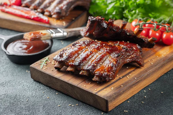 Primo piano di costolette di maiale grigliate con salsa barbecue e caramellate nel miele. Spuntino saporito a birra su un'asse di legno per limatura su sfondo concreto scuro — Foto Stock