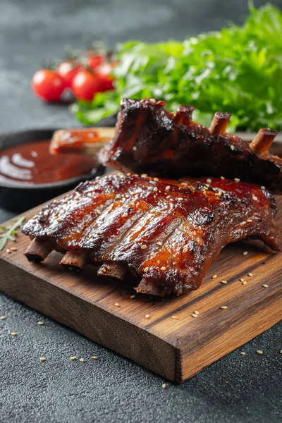 Primo piano di costolette di maiale grigliate con salsa barbecue e caramellate nel miele. Spuntino saporito a birra su un'asse di legno per limatura su sfondo concreto scuro — Foto Stock