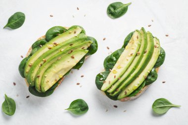 Dilimlenmiş avokado zeytin yağı, ıspanak ve keten ve susam tohumu ile iki ciabatta ekmeği. Sağlıklı vejetaryen kahvaltı beyaz arka plan üzerinde. Üstten görünüm. Düz yatıyordu