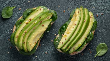 Dilimlenmiş avokado zeytin yağı, ıspanak ve keten ve susam tohumu ile iki ciabatta ekmeği. Sağlıklı vejetaryen kahvaltı karanlık bir arka plan üzerinde. Üstten görünüm. Düz yatıyordu