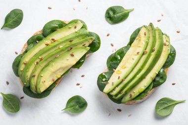Dilimlenmiş avokado zeytin yağı, ıspanak ve keten ve susam tohumu ile iki ciabatta ekmeği. Sağlıklı vejetaryen kahvaltı beyaz arka plan üzerinde. Üstten görünüm. Düz yatıyordu.