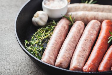 Karanlık bir beton masada kekik, biberiye ve dökme demir sarımsak ile çeşitli taze çiğ sosis tava.