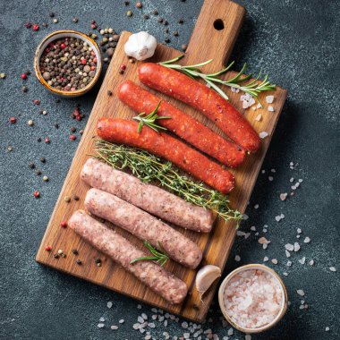 Kekik, biberiye, zeytinyağı ve sarımsak bir Ahşap Doğrama tahtası koyu bir beton tablo üzerinde çeşitli taze sosis. Siyah arka plan üzerine akşam yemeği için sosis pişirmeye hazır. Kopya alanı ile üstten görünüm.