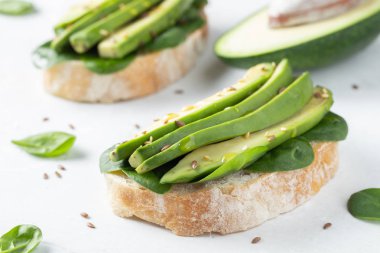 Dilimlenmiş avokado zeytin yağı, ıspanak ve keten ve susam tohumu ile iki ciabatta ekmeği. Sağlıklı vejetaryen kahvaltı beyaz arka plan üzerinde.