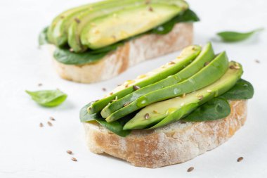 Dilimlenmiş avokado zeytin yağı, ıspanak ve keten ve susam tohumu ile iki ciabatta ekmeği. Sağlıklı vejetaryen kahvaltı beyaz arka plan üzerinde.