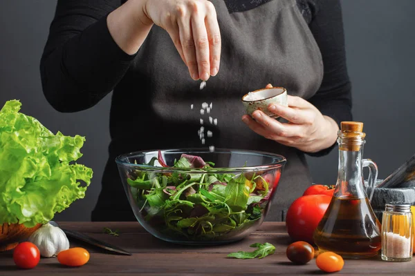 Woman chef in the kitchen preparing vegetable salad. Healthy Eating. Diet Concept. A Healthy Way Of Life. To Cook At Home. For Cooking. The girl sprinkles salt in a salad on a dark background.