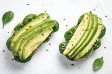 Dilimlenmiş avokado zeytin yağı, ıspanak ve keten ve susam tohumu ile iki ciabatta ekmeği. Sağlıklı vejetaryen kahvaltı beyaz arka plan üzerinde. Üstten görünüm. Düz yatıyordu