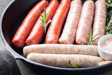Close-up assorted fresh raw sausages with thyme, rosemary and garlic in a cast iron pan on a dark concrete table