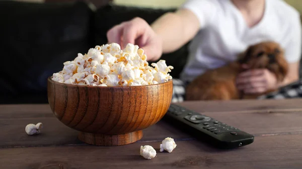 Wooden bowl with salted popcorn and TV remote on wooden table. In the background, a man with a red dog watching TV on the couch.