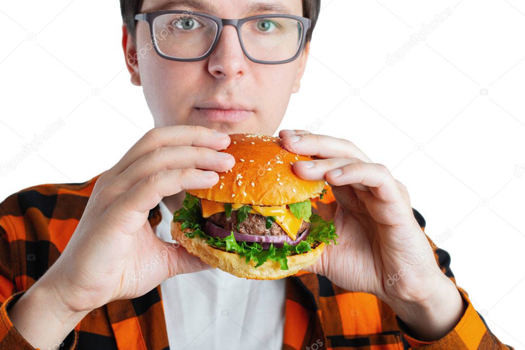 A young guy with glasses holding a fresh Burger. A very hungry student eats fast food. Hot helpful food. The concept of gluttony and unhealthy diet. With copy space for text. Isolated.