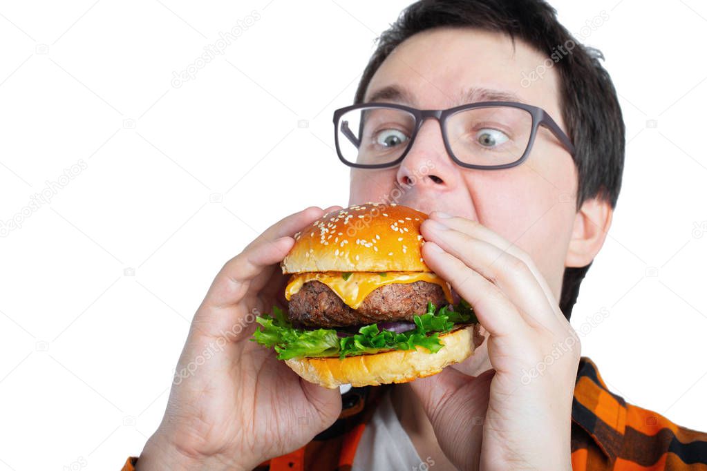 A young guy with glasses holding a fresh Burger. A very hungry student eats fast food. Hot helpful food. The concept of gluttony and unhealthy diet. With copy space for text. Isolated.