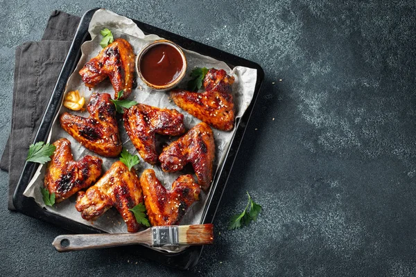 Roasted chicken wings in barbecue sauce with sesame seeds and parsley in a baking tray on a dark table. Top view with copy space. Tasty snack for beer on a dark background. Flat lay