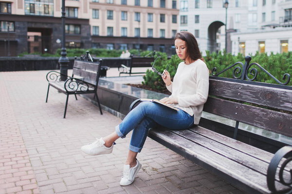 A beautiful young girl with long brown hair sitting on a bench with a book, holding eyeglasses. She left the house on a warm evening to read in the yard. The urban background.