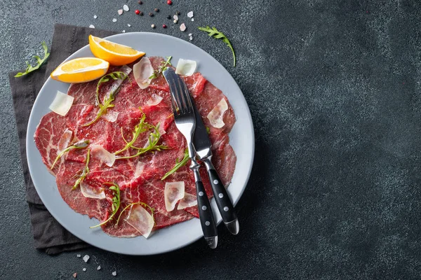 Marbled beef carpaccio with arugula, lemon and parmesan cheese on dark concrete table. Top view, flat lay with copy space — Stock Photo, Image
