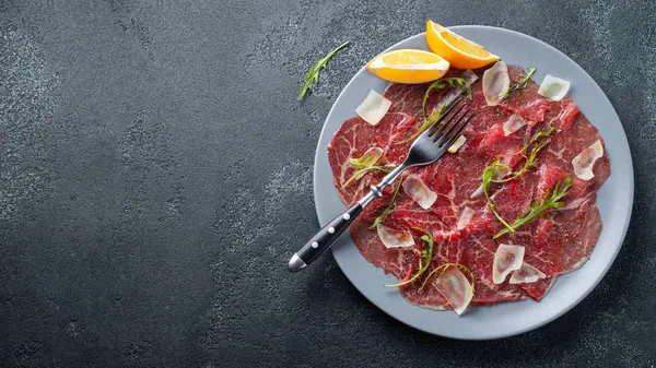 Gemarmerde Rundercarpaccio met rucola, citroen en Parmezaanse kaas op donkere betonnen tafel. Bovenaanzicht, vlakke lay met Kopieer ruimte — Stockfoto