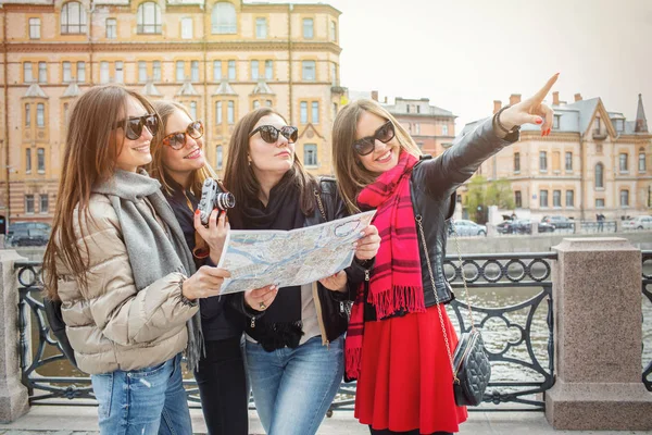 Un grupo de jóvenes turistas están buscando atracciones en una ciudad europea en el mapa. Cuatro mujeres alegres y hermosas caminan por la ciudad de primavera en gafas de sol . — Foto de Stock