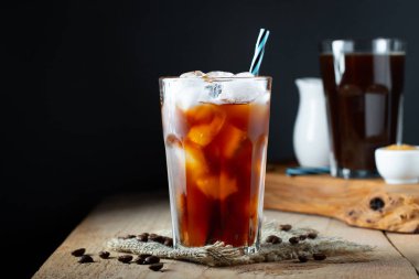 Krema ile uzun bir bardak buz kahve, eski bir rustik ahşap masa üzerinde buz küpleri ve fasulye dökülür. Kopya alanı ile siyah bir arka plan üzerinde tüpler ile soğuk yaz içkisi