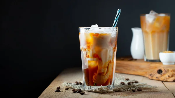 Eiskaffee in einem hohen Glas mit Sahne, Eiswürfeln und Bohnen auf einem alten rustikalen Holztisch. kaltes Sommergetränk mit Röhren auf schwarzem Hintergrund mit Kopierraum — Stockfoto