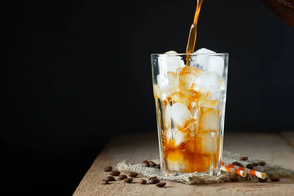 Es kopi dalam gelas tinggi dengan krim dituangkan di atas, es batu dan kacang-kacangan di atas meja kayu pedesaan tua. Minuman musim panas yang dingin dengan tabung di latar belakang hitam dengan ruang fotokopi — Stok Foto