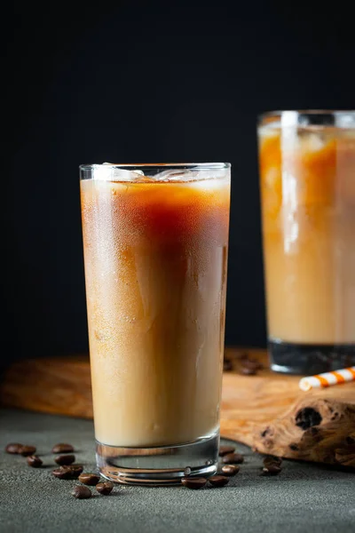 Es kopi dalam gelas tinggi dengan krim dituangkan di atas, es batu dan kacang-kacangan di atas meja kayu pedesaan tua. Minuman musim panas yang dingin dengan tabung di latar belakang hitam — Stok Foto