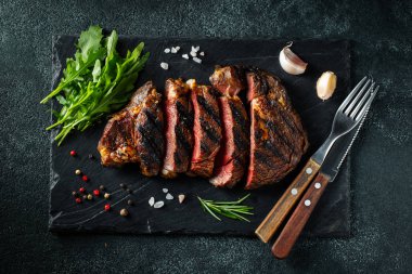 Dilimlenmiş biftek ribeye, biber, sarımsak, tuz ve kekik ile ızgara koyu taş arka plan üzerinde bir kayrak doğrama Kurulu servis edilir. Üst te. Düz yatıyordu