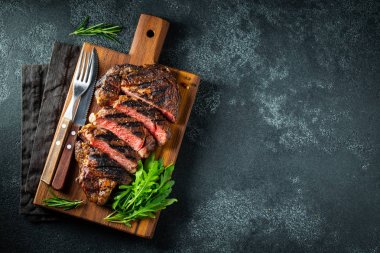 Dilimlenmiş biftek ribeye, biber ile ızgara, sarımsak, tuz ve keklik koyu taş arka planda bir tahta kesme kurulu üzerinde hizmet. Kopya alanı ile üst görünüm. Düz yatma