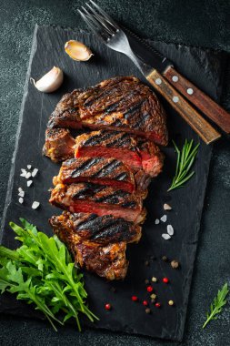Dilimlenmiş biftek ribeye, biber ile ızgara, sarımsak, tuz ve keklik koyu taş arka planda bir arduvaz kesme kurulu üzerinde hizmet. Üst görünüm. Düz yatma