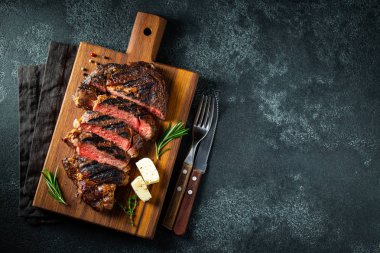 Dilimlenmiş biftek ribeye, biber ile ızgara, sarımsak, tuz ve keklik koyu taş arka planda bir tahta kesme kurulu üzerinde hizmet. Kopya alanı ile üst görünüm. Düz yatma