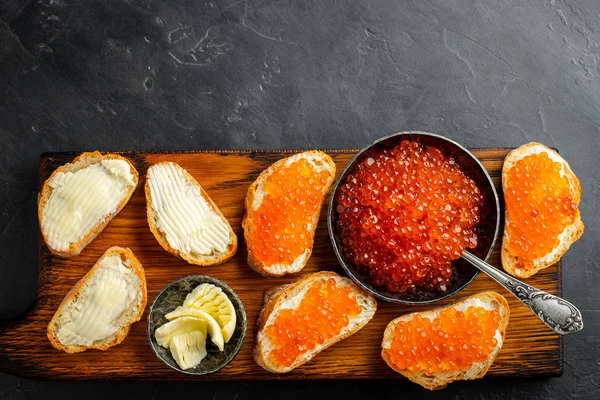 Close-up red caviar in bowl and Sandwiches on wooden cutting board on black background. Top view with copy space. Flat lay — Stock Photo, Image