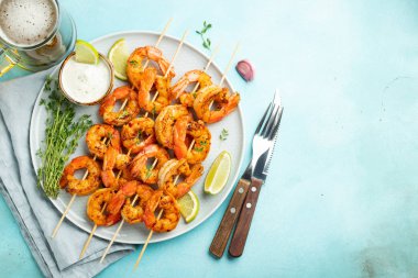 Açık mavi beton zemin üzerinde limon, sarımsak ve sosla servis edilen ızgara karides şişleri veya langoustinler. Deniz ürünleri ve bira. Kopyalama alanı olan üst görünüm. Düz yatıyordu