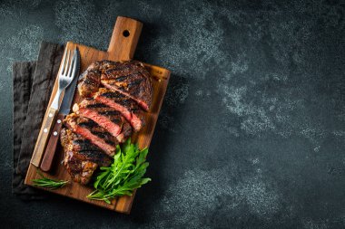 Dilimlenmiş biftek ribeye, biber ile ızgara, sarımsak, tuz ve keklik koyu taş arka planda bir tahta kesme kurulu üzerinde hizmet. Kopya alanı ile üst görünüm. Düz yatma