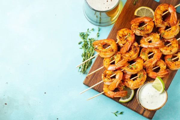 Spiedini di gamberi alla griglia o langoustine serviti con lime, aglio e salsa su uno sfondo di cemento azzurro. Pesce e birra. Vista dall'alto con spazio di copia. Posa piatta — Foto Stock