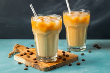 Büyük bardakta buzlu kahve, krema dökülmüş ve kahve çekirdekleri arka planda. Tüplerle soğuk yaz içkisi