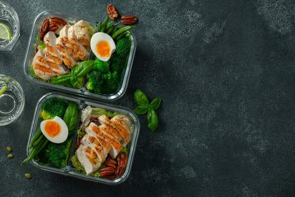 Healthy meal prep containers with green beans, chicken breast and broccoli. A set of food for keto diet in lunchbox on a dark concrete background. Top view with copy space.