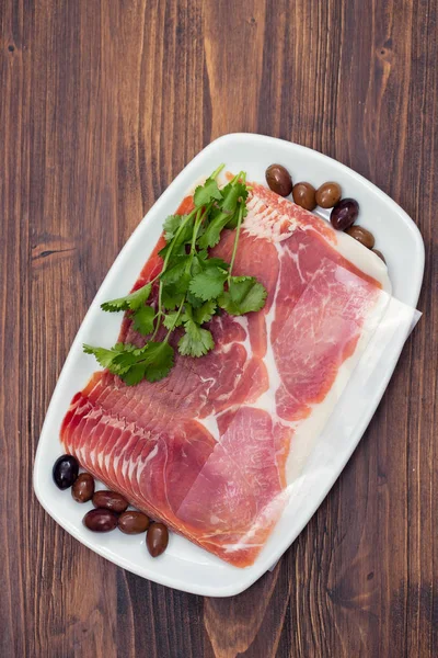 Prosciutto with olives and parsley on white dish on wooden backg — Stock Photo, Image