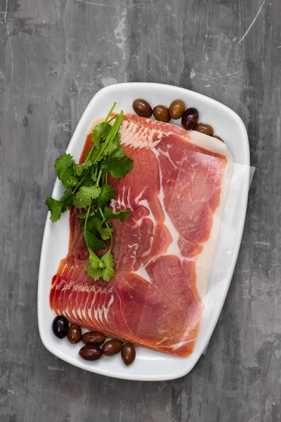 Prosciutto with olives and parsley on white dish on ceramic back — Stock Photo, Image