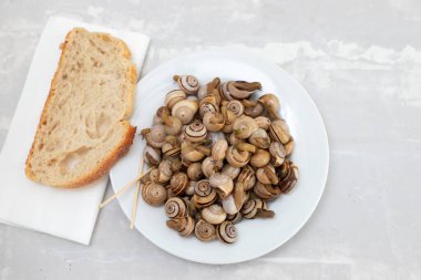 Beyaz tabakta kaynamış salyangoz ve seramik zemin üzerinde ekmek.