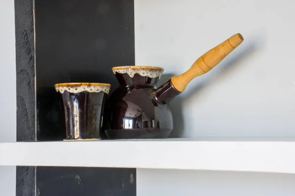 Ceramic cezve with wooden handle for brewing Turkish coffee and mug for coffee in brown standing on black and white shelf in kitchen room as the interior decor on the background of gray wall