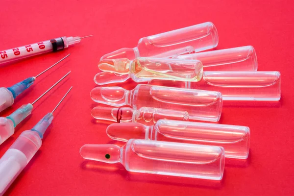 Pharmaceutical glass vials or ampules with liquid drug inside lie near syringes with needles on red uniform background. Concept preparation for treatment, course  injection therapy of various diseases
