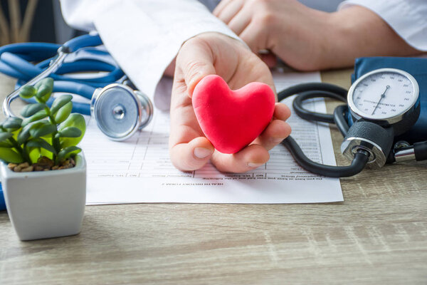 Doctor of internal medicine and cardiologist holding in his hands and shows to patient figure of red card heart during medical consultation. Explanation of causes of heart, diagnosis and treatment